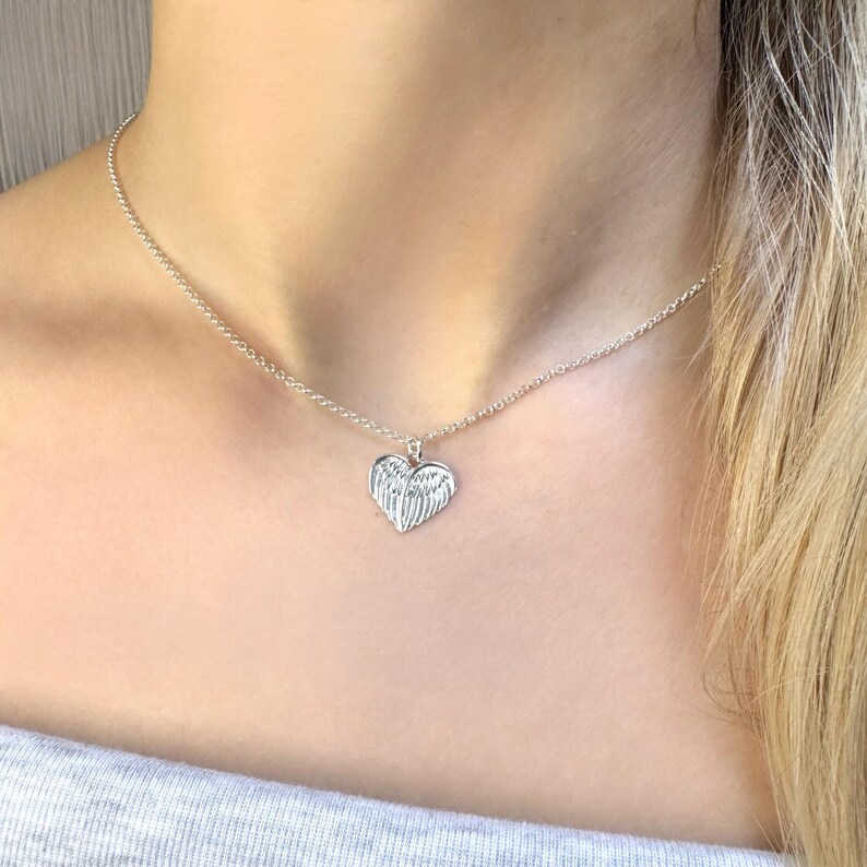 May include: A silver necklace featuring a heart-shaped pendant with wing details. The pendant hangs from a delicate chain. The necklace is worn around the neck.