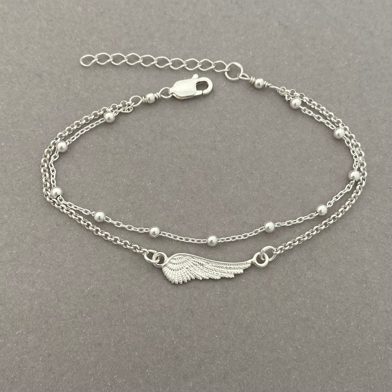 Puede incluir: Un brazalete de cadena de plata con un delicado dije de ala. El brazalete tiene un dise&ntilde;o de cadena doble con peque&ntilde;as cuentas.