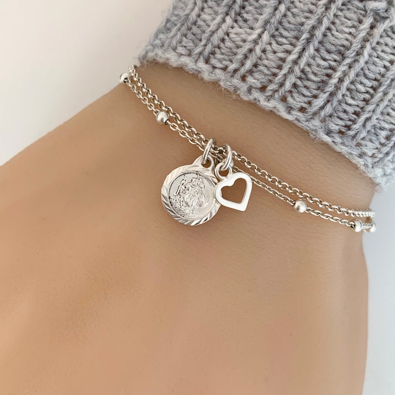 May include: A silver chain bracelet with a small heart charm and a round charm with a religious image. The bracelet is layered and has small silver beads.