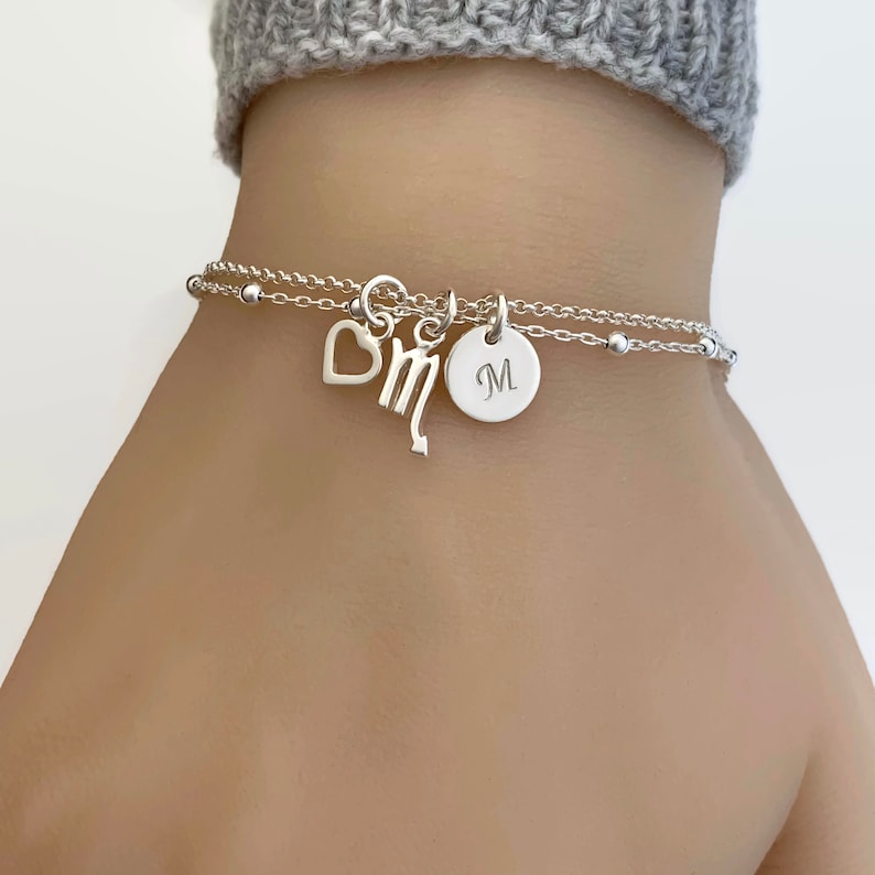 May include: A silver chain bracelet with a heart charm, a zodiac sign charm, and a personalized disc charm with the letter "M".