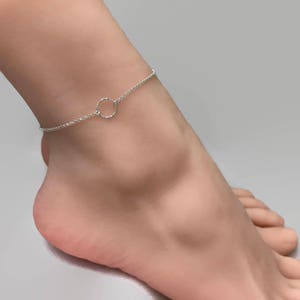 May include: A delicate silver anklet with a small open circle pendant. The anklet is worn on an ankle, showcasing the fine chain and minimalist design. The jewelry is a simple, elegant accessory.