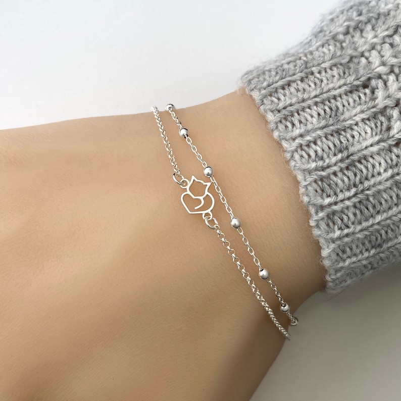 May include: A silver chain bracelet featuring a dainty charm of two cats cuddling. The bracelet is layered with a second silver chain with small beads.