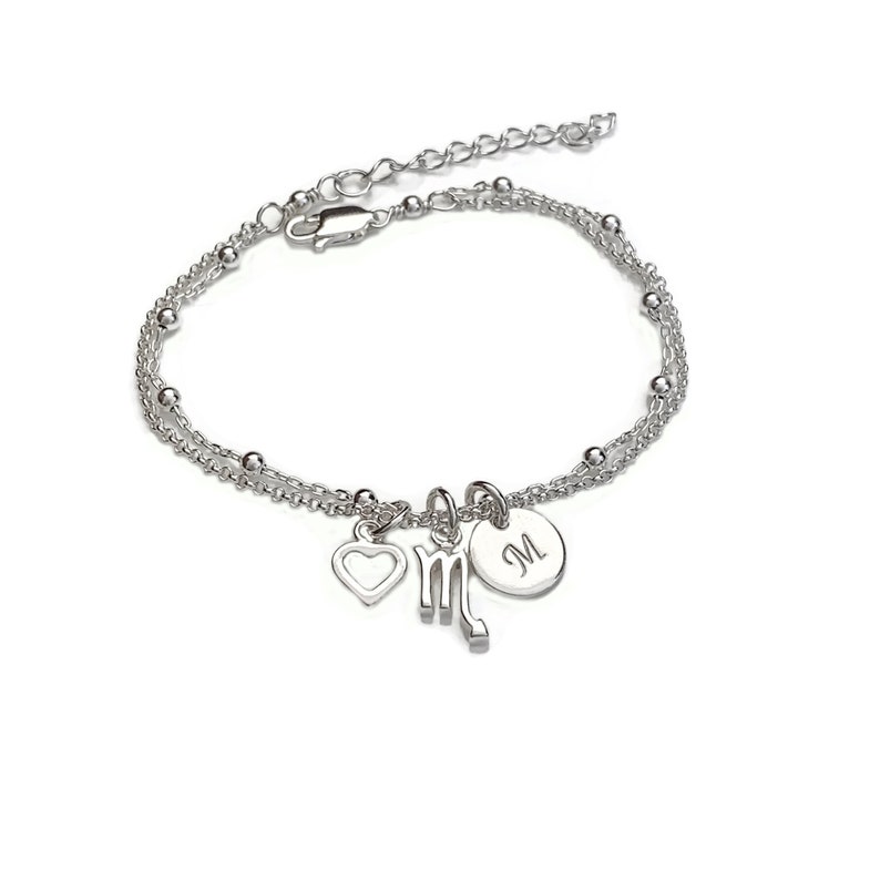 May include: Silver bracelet with a heart, a Scorpio zodiac sign charm, and a personalized initial charm. The initial charm is engraved with the letter "M".