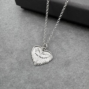 May include: A silver-colored necklace with a heart-shaped pendant featuring wing details. The pendant is suspended from a delicate chain. The necklace is displayed on a gray surface.