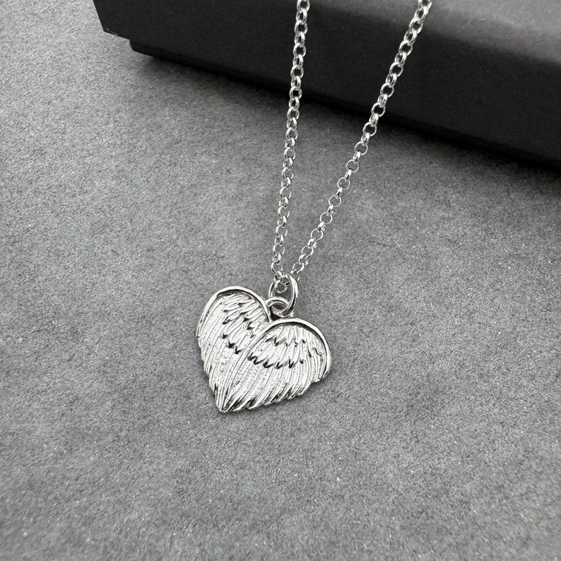 May include: A silver-colored necklace with a heart-shaped pendant featuring wing details. The pendant is suspended from a delicate chain. The necklace is displayed on a gray surface.