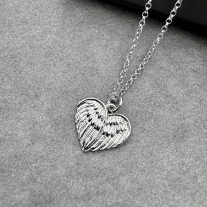 May include: A silver-colored necklace featuring a heart-shaped pendant with wing detailing. The pendant hangs from a delicate chain. The necklace is displayed on a gray surface.