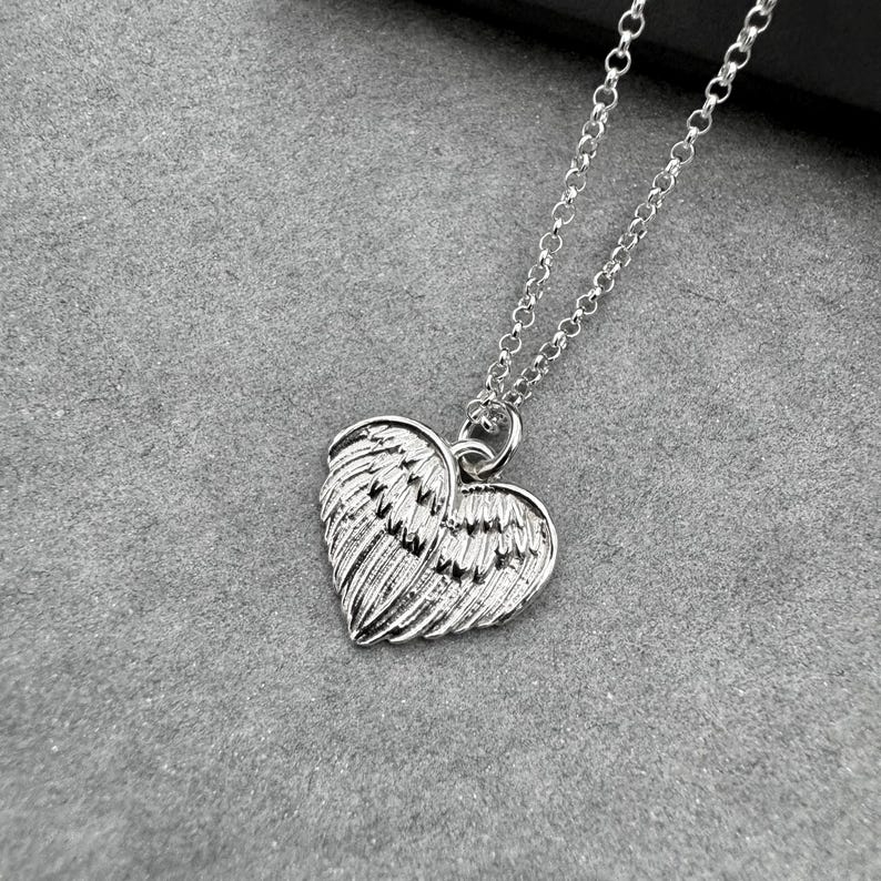 May include: A silver-colored necklace featuring a heart-shaped pendant with wing detailing. The pendant hangs from a delicate chain. The necklace is displayed on a gray surface.
