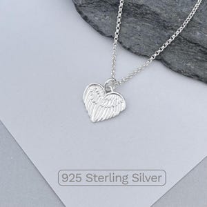 May include: A silver necklace with a heart-shaped pendant featuring wing details. The chain is delicate, and the pendant is made of 925 sterling silver. The necklace is displayed on a light gray surface.