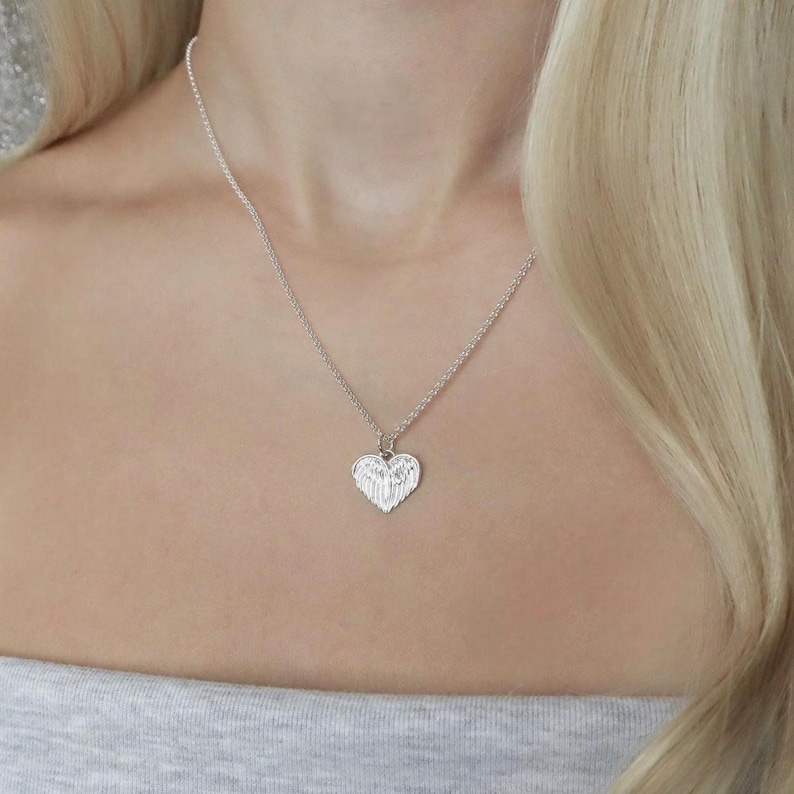 May include: A silver necklace featuring a heart-shaped pendant with an angel wing design. The pendant hangs from a delicate chain. The necklace is worn around the neck.
