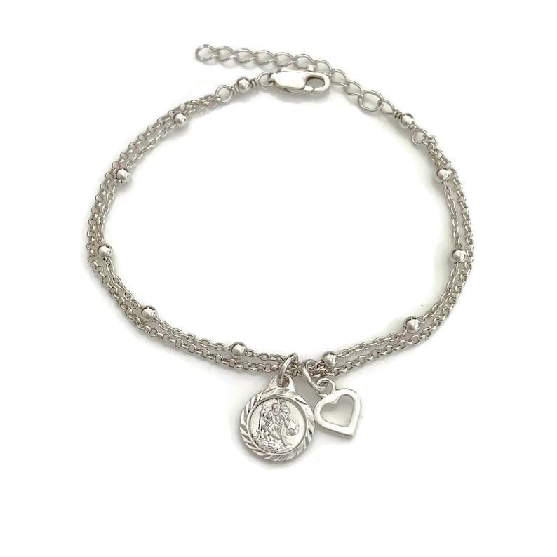 May include: A silver chain bracelet with a delicate heart charm and a round charm featuring a religious figure. The bracelet has a lobster clasp closure.