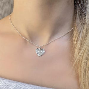 May include: A silver-colored necklace featuring a heart-shaped pendant with wing details. The pendant hangs from a delicate chain. The necklace is worn around the neck, with the pendant resting on the chest. The background is a blurred view of a person's upper body.