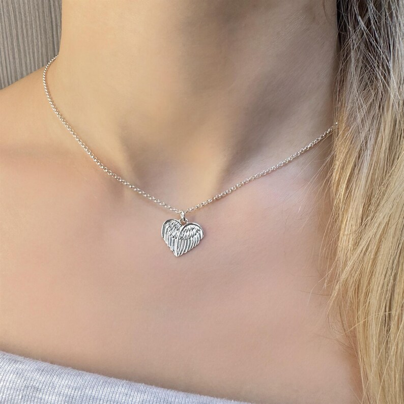 May include: A silver-colored necklace featuring a heart-shaped pendant with wing details. The pendant hangs from a delicate chain. The necklace is worn around the neck, with the pendant resting on the chest. The background is a blurred view of a person's upper body.