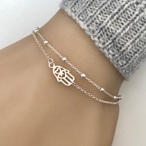 May include: A silver chain bracelet with a hamsa hand charm. The bracelet is layered with a second chain with small beads.
