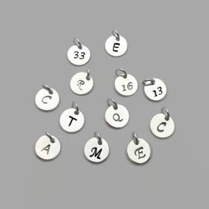 May include: A collection of silver charms with various letters and numbers engraved on them. The charms are round and have a hole at the top for attaching to a necklace or bracelet.