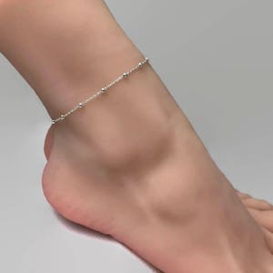 May include: A delicate silver anklet with small beads. The anklet is on a model's ankle.