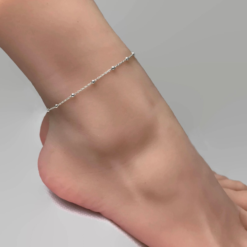 May include: A delicate silver anklet with small beads. The anklet is on a model's ankle.