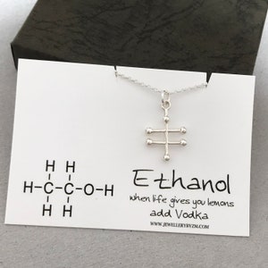 May include: A silver necklace with a pendant shaped like the chemical structure of ethanol. The pendant is on a white card with the text "Ethanol" and the chemical formula for ethanol. The card also says "when life gives you lemons add Vodka" and "WWW.JEWELLERYBYZM.COM".