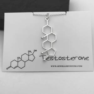 May include: A silver necklace with a pendant shaped like the chemical structure of testosterone. The pendant is on a white card with the word "Testosterone" written in black and the website "www.jewellerybyzm.com".