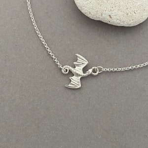 May include: A silver chain necklace with a small, detailed dragon pendant. The dragon is facing to the right and has its wings spread out.