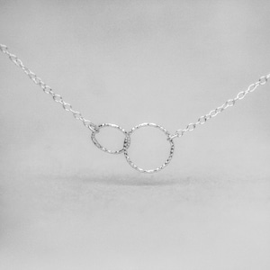 Peut inclure: Un collier argenté délicat avec deux cercles texturés entrelacés. Les cercles sont reliés par une fine chaîne, créant un design minimaliste et élégant. Le collier est présenté sur un fond doux et neutre.
