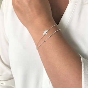 May include: A silver bracelet with a delicate chain and a small bird charm. The bracelet is layered with a second chain with tiny silver beads.
