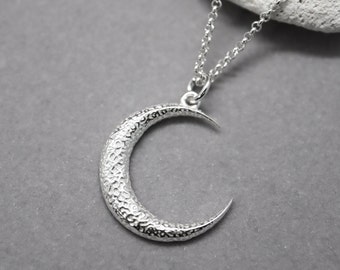 May include: A silver crescent moon necklace with a textured surface. The chain is silver and has a delicate design.