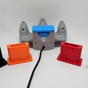 May include: A gray Nintendo 64 controller with a blue top and a black cord. Four different colored cartridge holders are displayed in front of the controller: black, orange, red, and white.