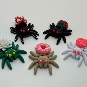 May include: Six small, colorful, 3D printed spiders with different toppings. The spiders are black, green, white, and brown. The toppings include a mushroom, a strawberry, a flower, a donut, and a rose.