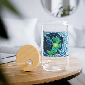 May include: A clear glass can-shaped drinking glass with a bamboo lid and straw. The glass features a colorful abstract design with blue, green, and white hues. The lid and straw are made of natural bamboo.