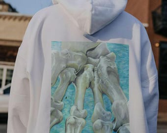 Puede incluir: Sudadera blanca con una impresión gráfica de un esqueleto de mano en la espalda. El esqueleto está dibujado en un estilo realista con huesos y articulaciones detallados. El fondo de la impresión es una acuarela azul claro.