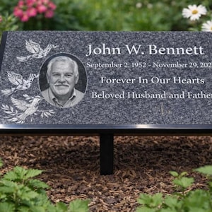 May include: A rectangular memorial plaque with a black border, set in a garden. The plaque features an image of a man, the name "John W. Bennett", dates, and the inscription "Forever In Our Hearts, Beloved Husband and Father". Decorative doves are etched on the plaque.