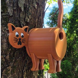 May include: A brown wooden birdhouse shaped like a cat with a hole for birds to enter. The cat has a brown body, a black nose, and white eyes. The birdhouse is mounted on a tree trunk.
