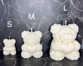 Pode incluir: Três velas de urso de rosa branca em tamanhos diferentes, etiquetadas como S, M e L. As velas são feitas de cera branca e têm um design em forma de coração no centro.