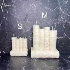 May include: Two white candle sets shaped like stacks of books. The smaller set is labelled "S" and the larger set is labelled "M".
