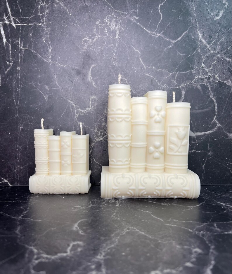May include: Two sets of white pillar candles with decorative designs. The larger set has four candles and the smaller set has three candles. Both sets are arranged in a stack and have a base that looks like a book.