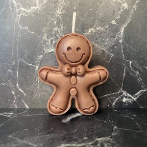 Könnte beinhalten: Eine braune Lebkuchenmann-Kerze mit einem lächelnden Gesicht, einer Fliege und Knopfformen. Die Kerze hat einen Docht oben. Sie steht vor einem dunklen, marmorierten Hintergrund. Festliche Heimdekoration.