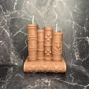 May include: A set of three brown decorative candles shaped like books with intricate designs. The candles are arranged in a stack with the bottom candle serving as a base. Each candle has a white wick.