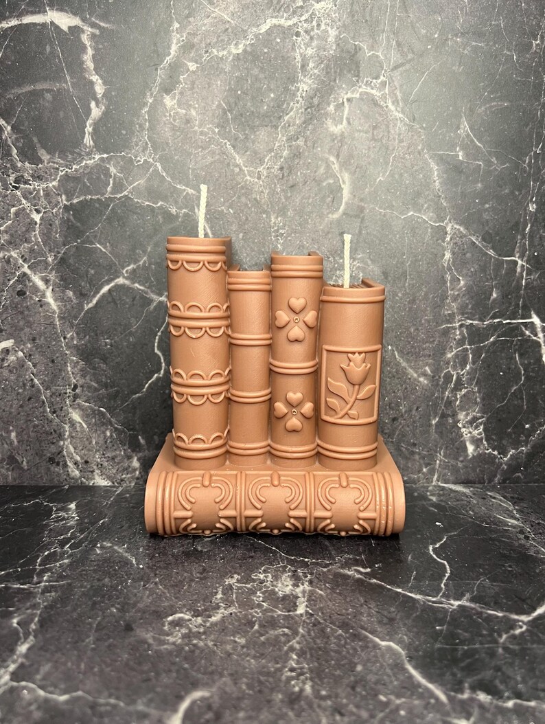 May include: A set of three brown decorative candles shaped like books with intricate designs. The candles are arranged in a stack with the bottom candle serving as a base. Each candle has a white wick.