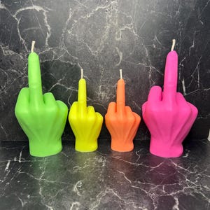 Könnte beinhalten: Vier farbige Kerzen in Form einer Hand, die den Mittelfinger zeigt. Die Kerzen sind neongrün, gelb, orange und pink. Jede Kerze hat einen weißen Docht. Sie stehen auf einer dunklen, marmorierten Oberfläche.