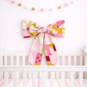 May include: A large, patchwork fabric bow in shades of pink, yellow, and burgundy hangs above a white crib. The bow is made of various patterned fabrics. A garland of pink and gold felt balls decorates the wall. The crib has a striped pink and yellow sheet.