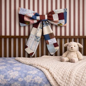 May include: A nursery scene featuring a wooden crib with a blue floral patterned sheet and a cream-colored knitted blanket. A stuffed animal dog sits on the blanket. A large patchwork bow hangs on the striped wall above the crib.