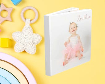 May include: A white photo book with a picture of a baby girl wearing a pink tutu. The book is next to a white crocheted star, wooden toys, and a rainbow-colored arch on a yellow background. The text "Zoe Mia" is on the book.