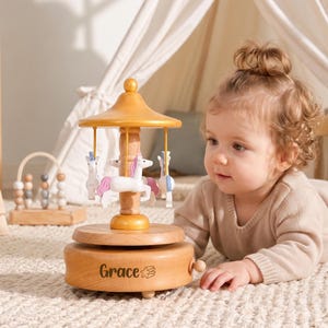 May include: A wooden carousel music box featuring white and pink unicorn figures suspended from a gold-toned canopy. The circular base is crafted from light-toned wood and features the name Grace engraved in black script with a small star icon.