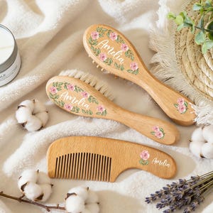 May include: Two wooden hairbrushes with soft bristles and one wooden comb, all featuring a floral design of pink roses and green leaves with the name Amelia printed in white script. The items have a light wood grain finish and floral accents.