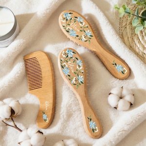 Op de afbeelding: Houten haarborstels en een kam met blauwe en witte bloemmotieven. De naam "Olivia" is op de items gegraveerd. Ze liggen op een witte, zachte ondergrond, samen met katoentakjes en een kaars.