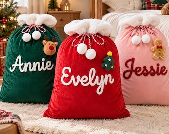 Pode incluir: Três sacos de presente de Natal de pelúcia nas cores verde escuro, vermelho e rosa claro. Cada saco possui uma borda de pele sintética branca, um cordão com pompons e um nome bordado em letra cursiva: Annie, Evelyn e Jessie.