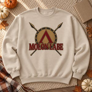 May include: A beige crewneck sweatshirt featuring a graphic print of a gold circular shield with a red Greek letter Lambda, crossed by two spears. The text MOLON LABE is printed in red with a dark outline across the centre of the design.