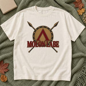 Puede incluir: Una camiseta de manga corta de color crema con un gráfico de un escudo circular de bronce con un símbolo lambda espartano de color rojo oscuro. Dos lanzas cruzadas se sitúan detrás del escudo y el texto MOLON LABE aparece impreso en rojo.