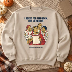 May include: A beige crewneck sweatshirt featuring a graphic illustration of Julius Caesar holding a scroll, surrounded by four figures in Roman attire. The text reads: I ASKED FOR FEEDBACK. NOT 23 POINTS. Julius Caesar • 44 BC in dark blue lettering.