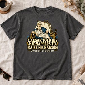 May include: A charcoal gray t-shirt featuring a graphic of a Roman figure on a ship with the text: CAESAR TOLD HIS KIDNAPPERS TO RAISE HIS RANSOM 20 talents? I'm worth 50. The design includes a white sail, blue cloak, and gold laurel accents.